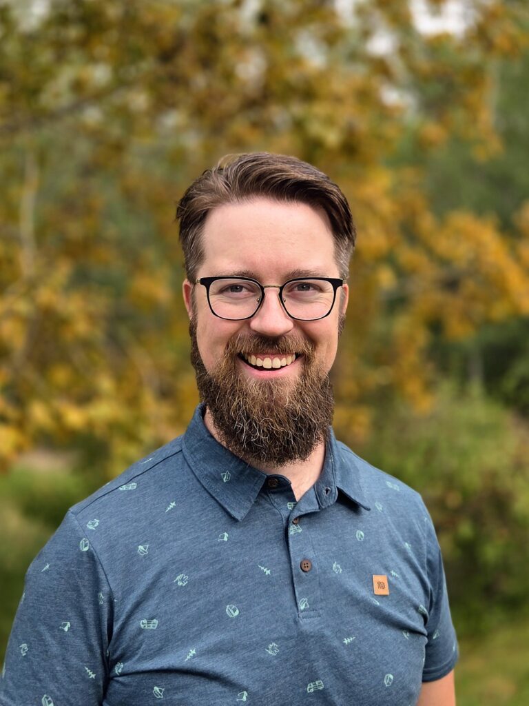 man with blue collared shirt and glasses, short brown hair and a large beard, in front of blurry autumn forest scenery.
